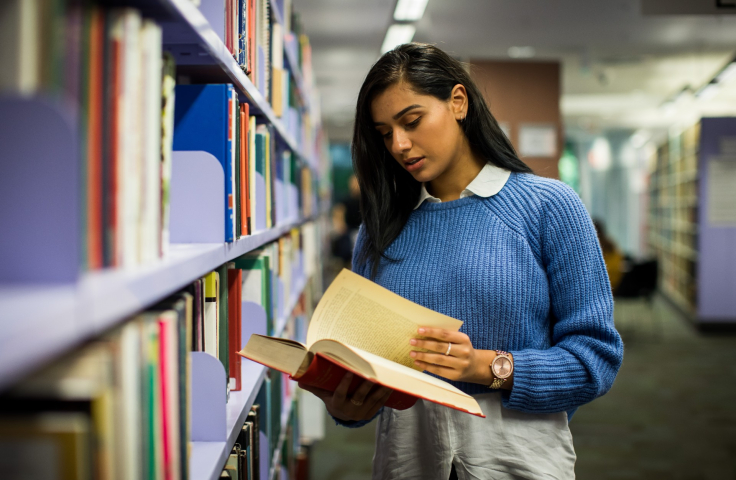 A woman with long back hair and a blue jumper reads a book in library
