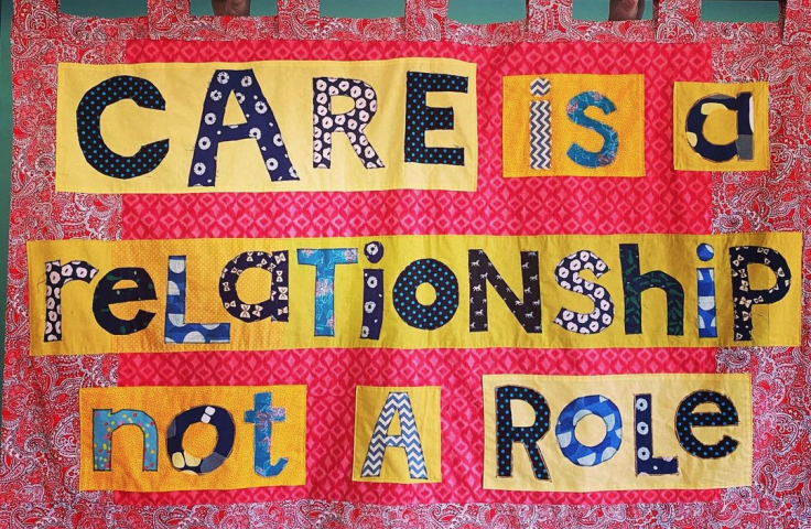 A handmade pink material banner held up by a curtain pole with the words 'Care is a Relationship not a Role' patchworked on to a yellow background