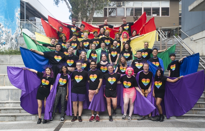 A number of people are standing in UNSW Pride shirts and holding flags that make up a rainbow