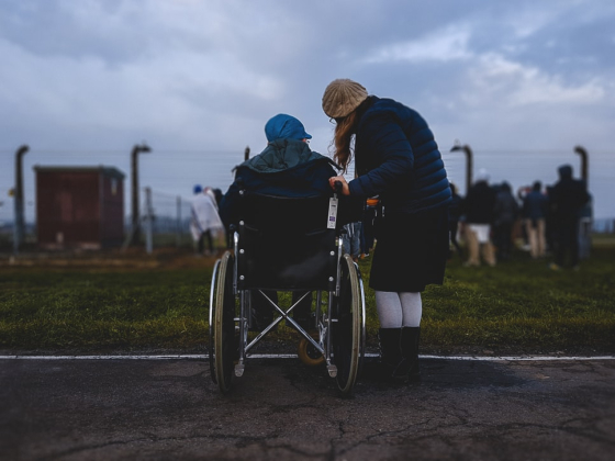 Outside on a pathway, looking at the back of someone in a wheelchair being supported by a female