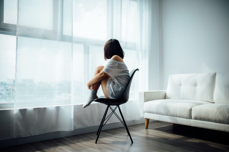 Woman in a t'shirt sat on a chair in an empty room looking out the window