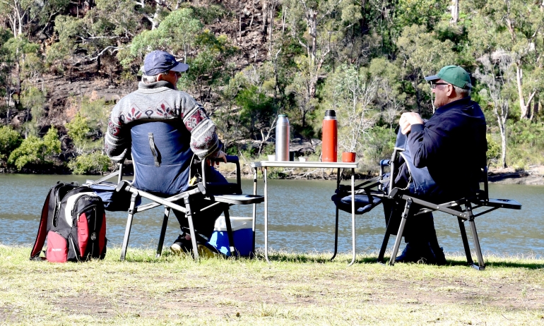 Two elder men sat in camp chairs by a river fishing with flasks  of hot drinks