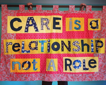 A handmade pink material banner held up by a curtain pole with the words 'Care is a Relationship not a Role' patchworked on to a yellow background
