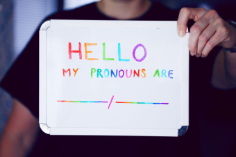 Person holding up a whiteboard with the words "Hello, my pronouns are..." in rainbow marker