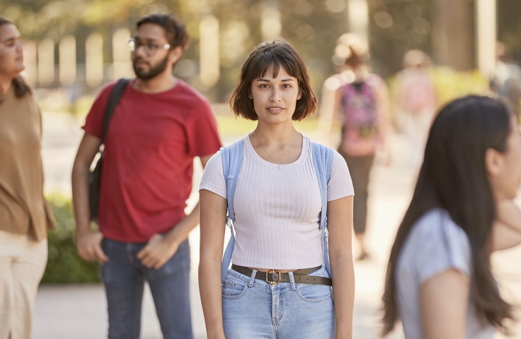 Female with cropped brown hair and a white t'shirt and jeans looking at camera with other students milling in the background of UNSW campus