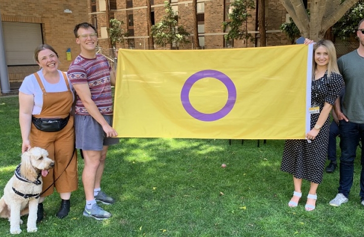 Four people holding the Intersex Flag and smiling towards the camera. From left Bethany Ross (and her dog Eddie), Mitchell McBurnie, Jessica Luquin and Michael Ormsby.  