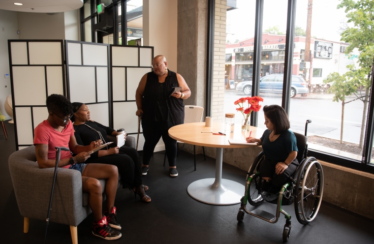 Four disabled people of color face each other in a open circle during a meeting. Two Black people sit on a couch with a cane leaning off the side while a Black non-binary person stands with a tablet and cane. A South Asian person in a wheelchair takes notes.