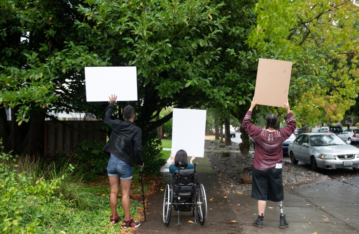 Three disabled people of color (a Black non-binary person with a cane, a South Asian person in a wheelchair, and an Indigenous Two-Spirit person with a prosthetic leg) block a neighborhood street while holding up cardboard signs. The photo is shot from behind everyone.