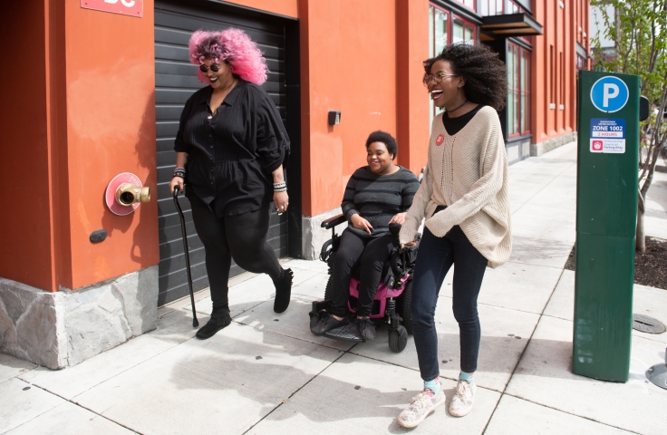 Three Black and disabled folx laugh uproariously while strolling down a sidewalk on a windy day. On the left, a non-binary person walks with a cane in one hand and a tangle stim toy in the other. In the middle, a non-binary person rolls along in their power wheelchair. On the right, a femme walks with fabulously windswept hair. A street parking meter is in the background on the right.