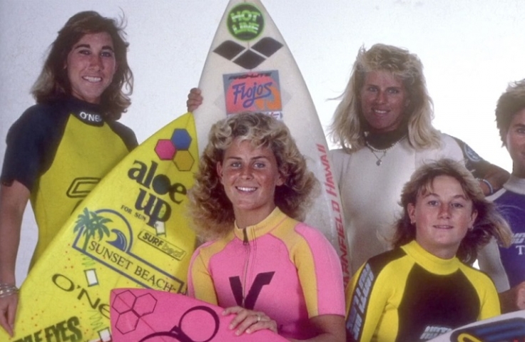 Five female surfers holding surfboards and smiling. 