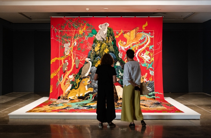 Two people standing in front of a tapestry made by Khadmin Ali. 
