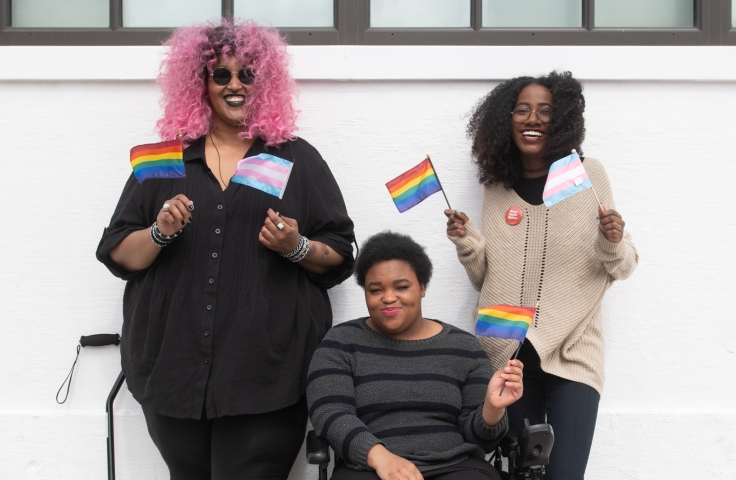 Three Black and disabled folx smiling and displaying mini flags - Disabled And Here