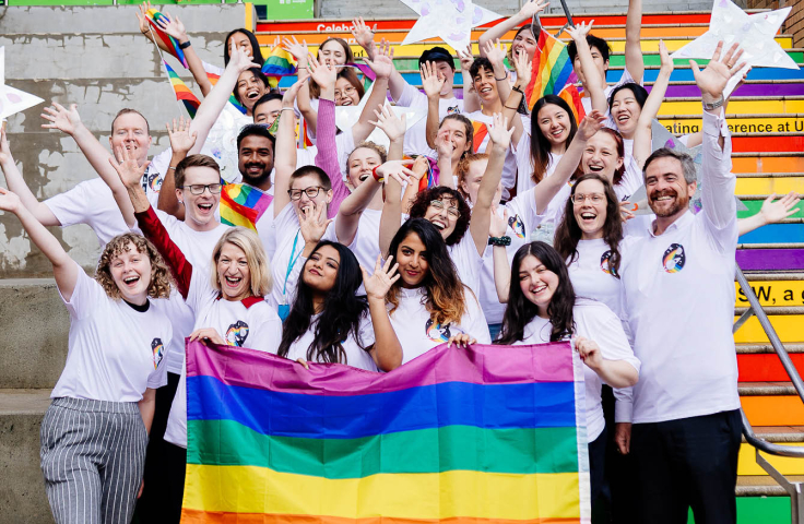 Group of staff and students on rainbow steps with rainbow flag and arms up