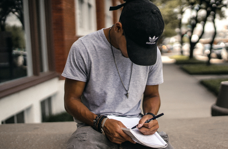 A young black man in a black baseball cap, grey shirt and jeans sits on a wall with his head down, writing on a pad