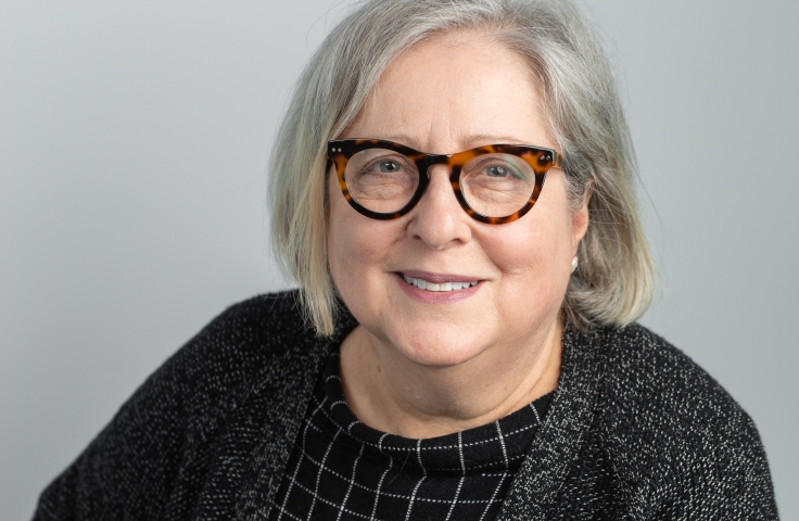 Betsy has shoulder length grey hair, glasses and wears a black check top and grey cardigan
