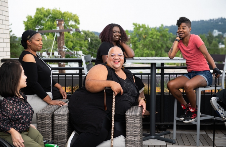 A Black non-binary person laughs while sitting in a cushioned wicker chair with their leopard print cane. They are surrounded by four more disabled people of color. Everyone is in the midst of conversation at a rooftop deck party - Disabled and Here