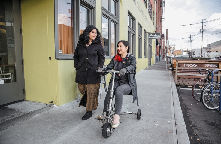 A Latinx (invisibly) disabled woman talking and walking alongside her friend, an Asian disabled woman wearing compression gloves and driving a lightweight electric mobility scooter - Disabled And Here