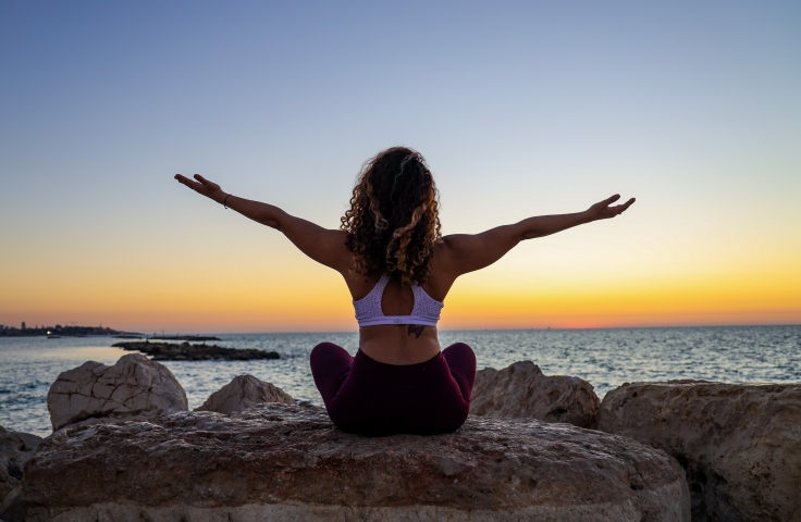 Woman sat with her back to the camera doing yoga pose on rocks at sunrise