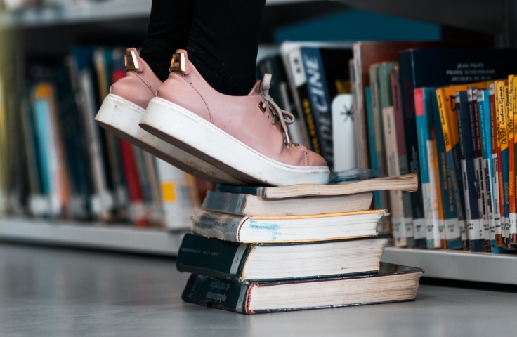 Person with pink trainers standing on books by a book shelf