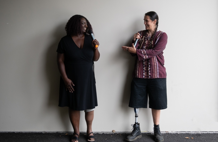 An Indigenous Two-Spirit person and a Black woman face each other and gesture while smiling and speaking into microphones - Disabled And Here