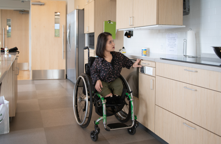 A South Asian person sits in her wheelchair in an accessible kitchen - Disabled And Here.